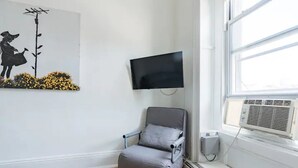Una habitación con un sillón gris, un televisor de pantalla plana montado en la pared y una ventana con un equipo de aire acondicionado.