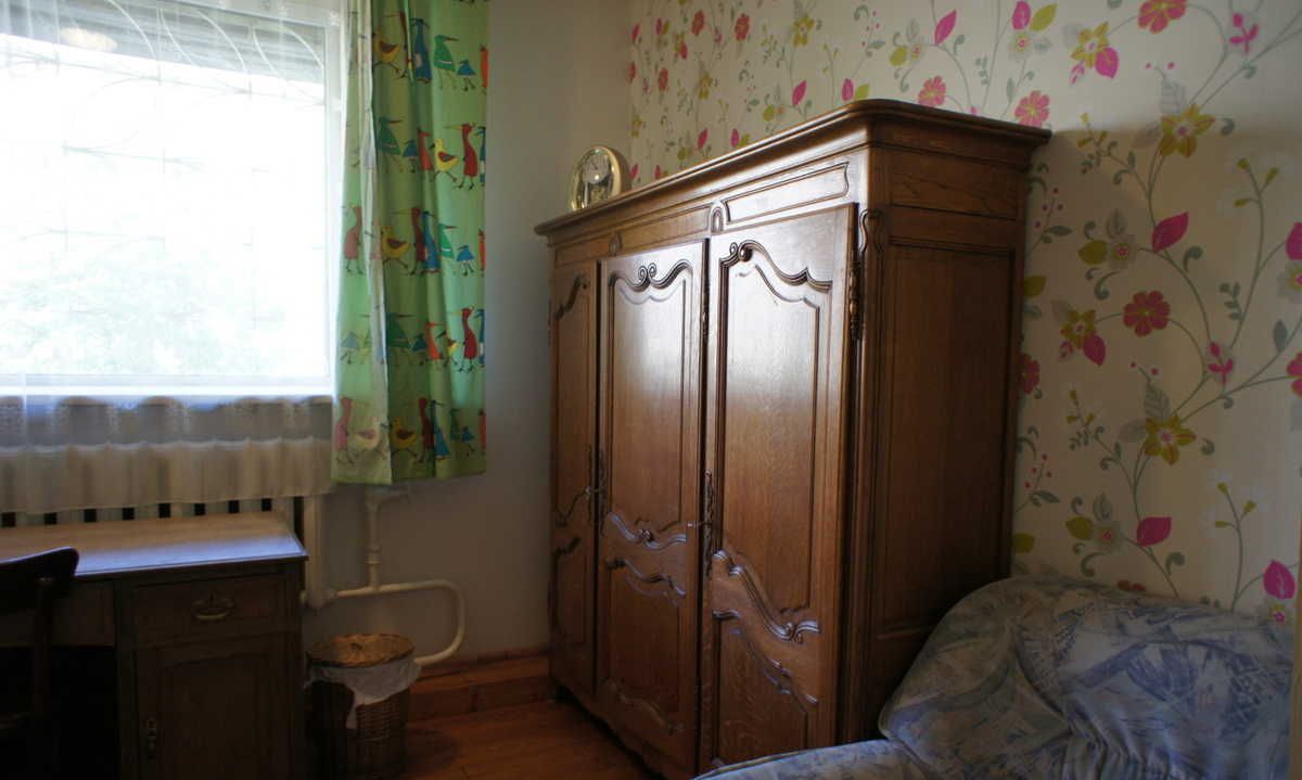 Une pièce avec une armoire en bois, un lit recouvert d’une couverture à motifs, une fenêtre avec des rideaux et un radiateur.