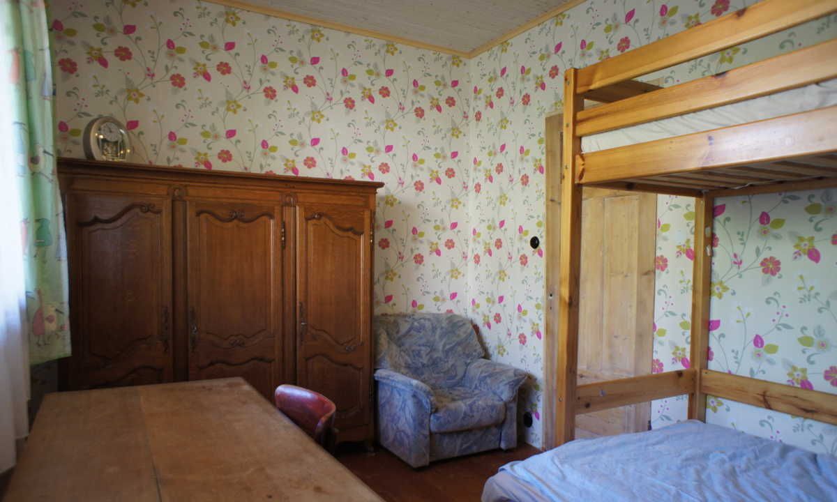 A room with a wooden bunk bed, a wooden wardrobe, a floral-patterned chair, and a table.