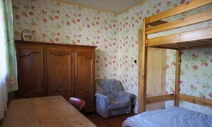 Una habitación con una cama litera de madera, un armario de madera, una silla estampada con motivos florales y una mesa.