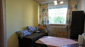 Une petite pièce agréable et confortable, avec un lit, une table et une chaise. Il y a une fenêtre avec des rideaux, un radiateur et une porte.