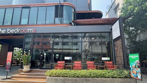 A modern building with a glass facade, a sign reading "the bedrooms", and outdoor seating with red chairs. - The Bedrooms Maeklong Service Apartment (Samut Songkhram)
