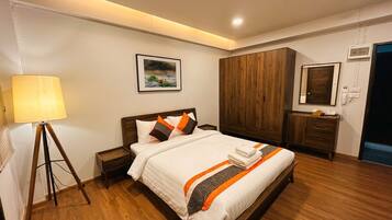 A hotel room with a bed, bedside table, lamp, wardrobe, and a painting on the wall.