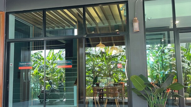 A modern building with large glass doors, a sign for 'The Bedrooms' hotel, and a potted plant in the foreground. - The Bedrooms Maeklong Service Apartment (Samut Songkhram)
