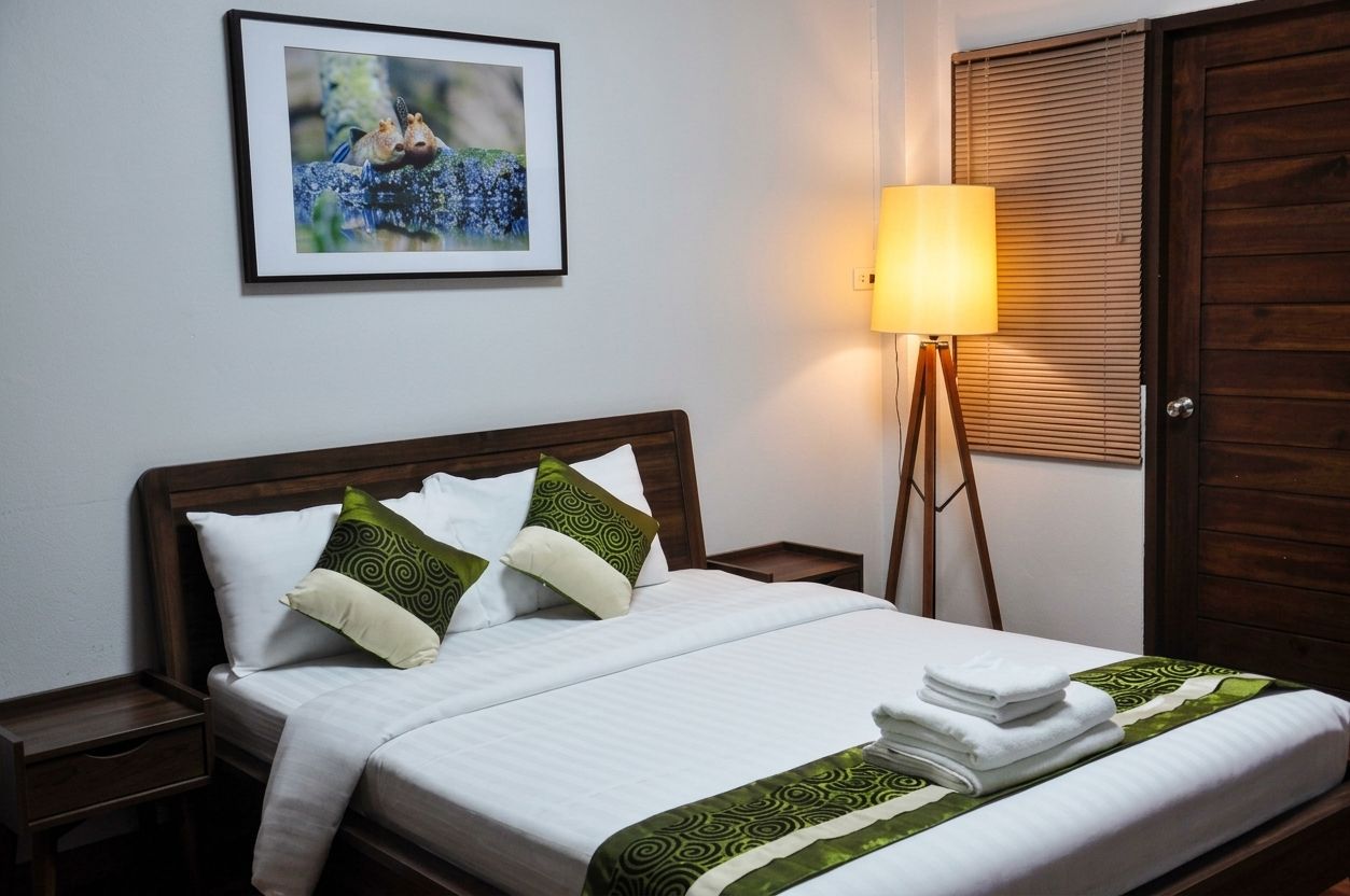 Una cama bien hecha con ropa de cama blanca y almohadas estampadas en verde, una lámpara de noche y un cuadro en la pared.
