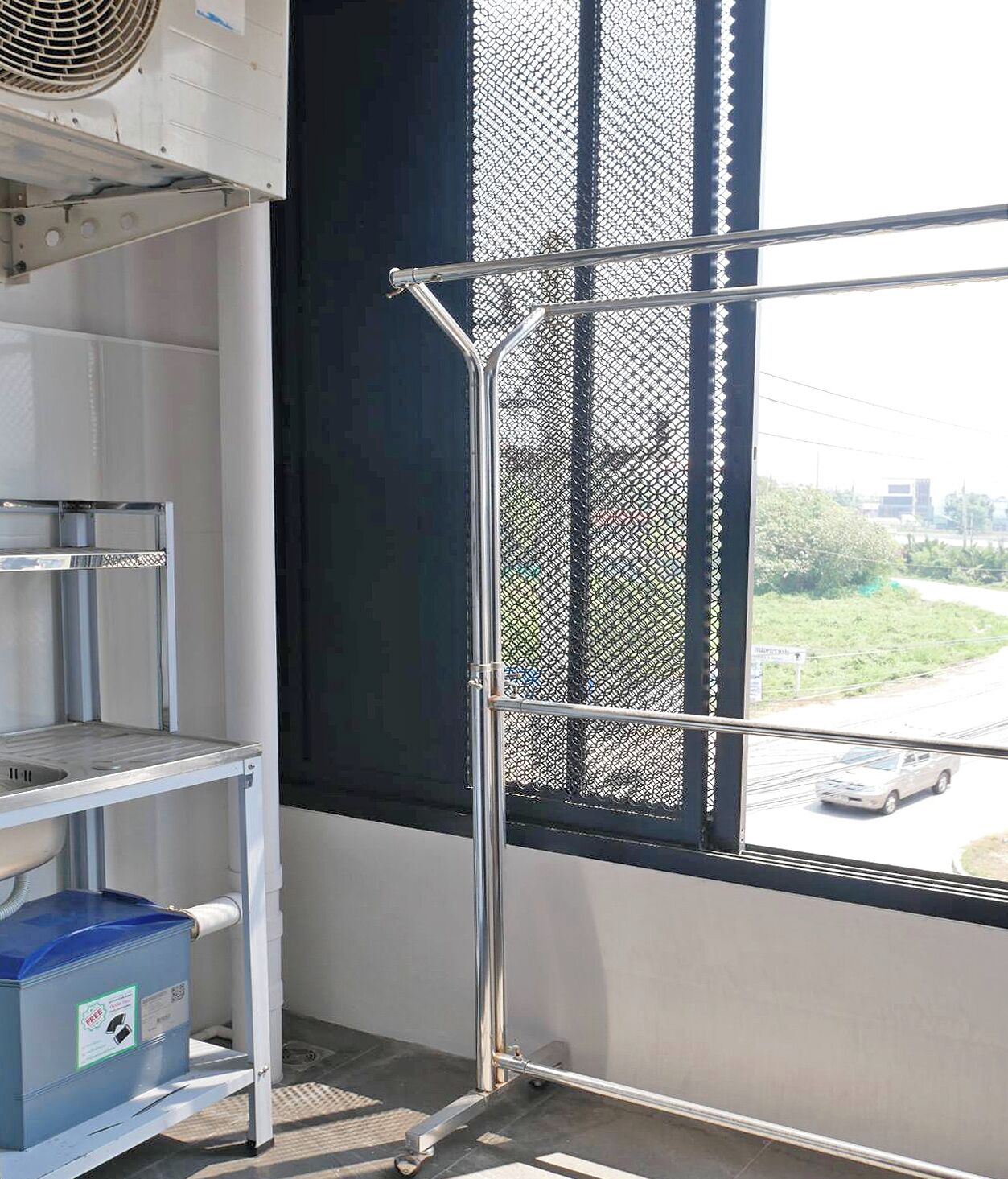 Esquina de una habitación con un estante metálico, un recipiente azul y una ventana que da a la calle.