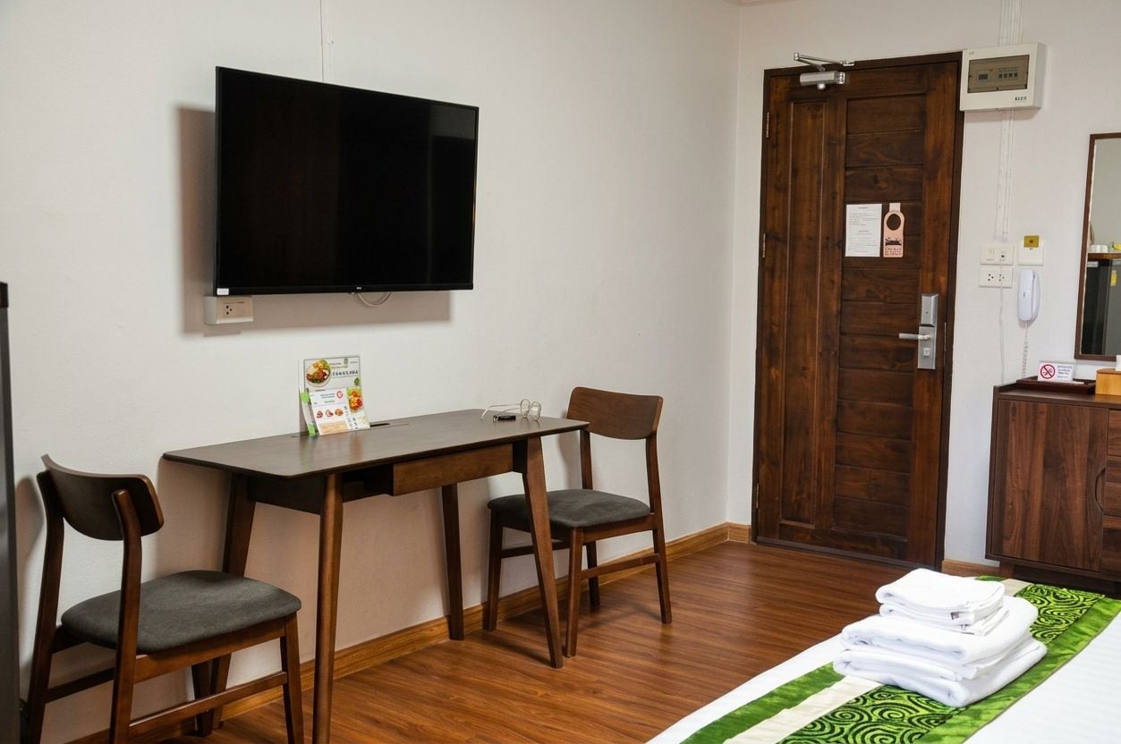 A hotel room with a flat-screen TV mounted on the wall, a wooden desk with two chairs, a wooden door, a small cabinet with a mirror, and a neatly folded stack of towels on the floor.