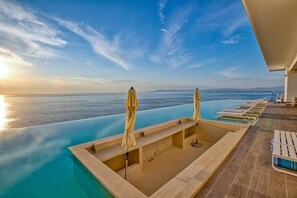 Una piscina infinita con sillas de descanso y sombrillas, con vista al mar tranquilo y a la costa lejana.