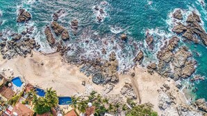 An aerial view of a beach with rocky formations, a swimming pool, and a few buildings.