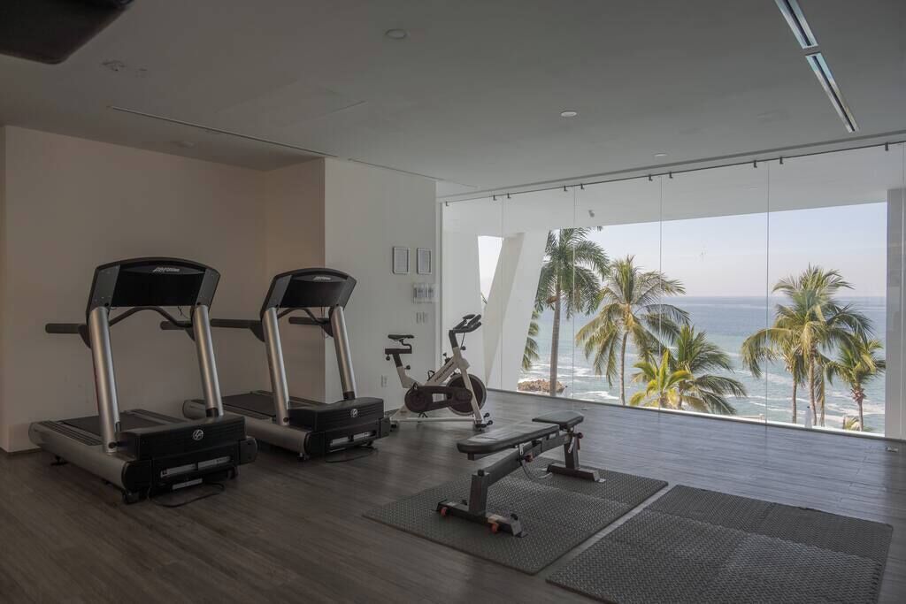Een sportschool met loopbanden, een elliptische trainer en een bank, met uitzicht op een strand met palmbomen.