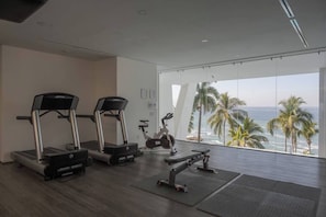 Ein Fitnessraum mit Laufbändern, Crosstrainern und einer Bank, mit Blick auf einen Strand mit Palmen.