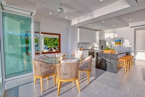 Una cocina moderna con una isla central, un comedor con sillas de mimbre y un amplio ventanal que da a un espacio exterior.