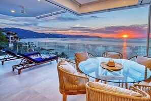 Een balkon met een glazen tafel, rieten stoelen en ligstoelen met uitzicht op een zonsondergang boven de zee.