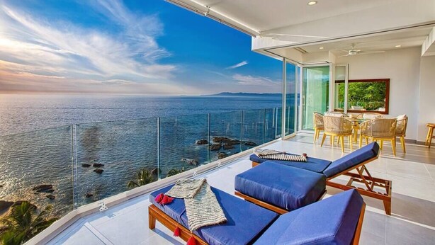 A balcony with a view of the sea, two lounge chairs, a dining table with chairs, and a ceiling fan.