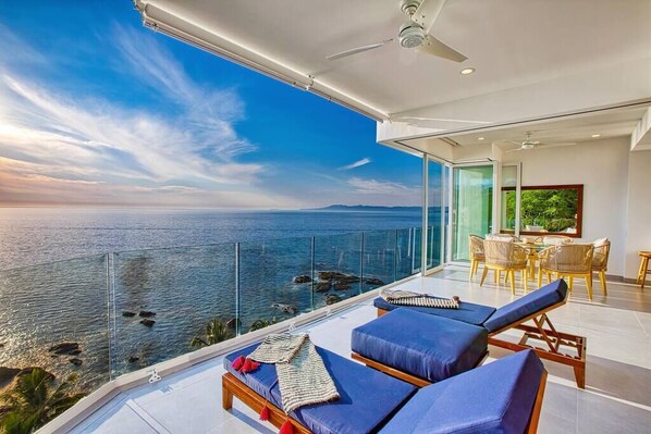 A balcony with a view of the sea, two lounge chairs, a dining table with chairs, and a ceiling fan.