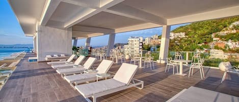 A rooftop lounge with sun loungers and tables, offering a view of the city and the sea.