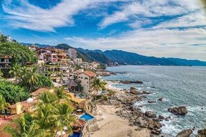 Un pueblo costero con edificios a lo largo de la playa, un litoral rocoso y montañas al fondo.