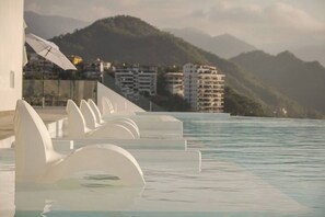 A poolside area with white lounge chairs, a glass railing, and a view of buildings and mountains in the background.