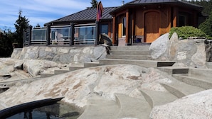 Una casa moderna con una escalera de piedra que conduce a una puerta de madera, rodeada de grandes rocas y un pequeño estanque.