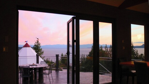 Habitación con puerta corrediza de vidrio que da a una terraza exterior con mesa y sillas, con vista a un cuerpo de agua y montañas.