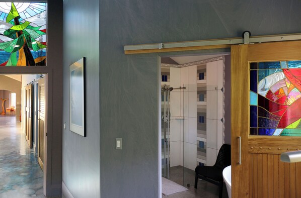 Un pasillo con una puerta de vidrio que da a la zona de la ducha, una puerta de madera con un vitral y un cuadro en la pared.