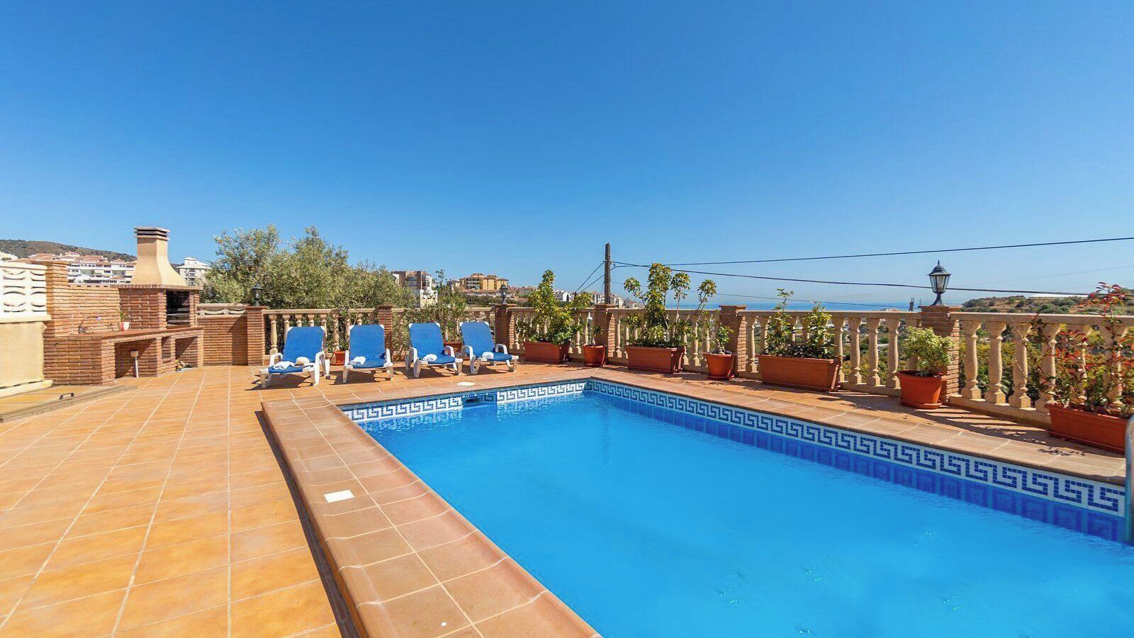 A rooftop swimming pool with blue tiles, lounge chairs, and potted plants.
