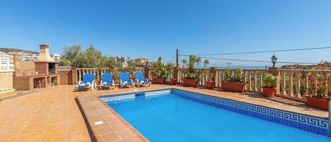 A rooftop swimming pool with blue tiles, lounge chairs, and potted plants.