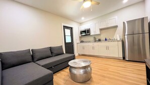A modern living room with a grey sectional sofa, a round metallic coffee table, a ceiling fan, and a kitchen area with white cabinets and stainless steel appliances. - The Romantic Bird House | A Storybook Garden Retreat (Homestead)