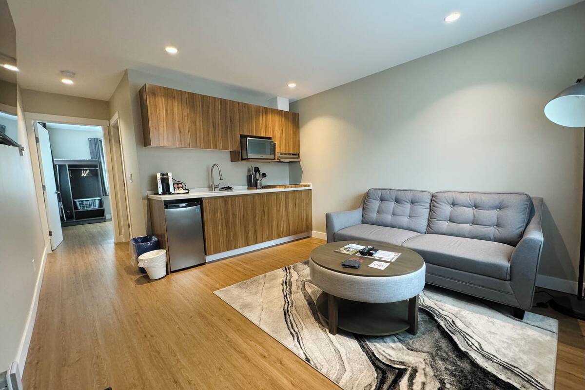 A modern living room with a grey sofa, a round coffee table, and a kitchen area with wooden cabinets and a built-in microwave.