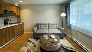 A modern living room with a grey sofa, a small round ottoman, a coffee table with remote controls and magazines, a kitchen area with wooden cabinets and a microwave, and a large window with sheer curtains. - Kitimat River Motel & Suites (Kitimat)