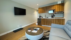 A modern living room with a grey sofa, a round coffee table, and a kitchen area with wooden cabinets and a built-in microwave. - Kitimat River Motel & Suites (Kitimat)