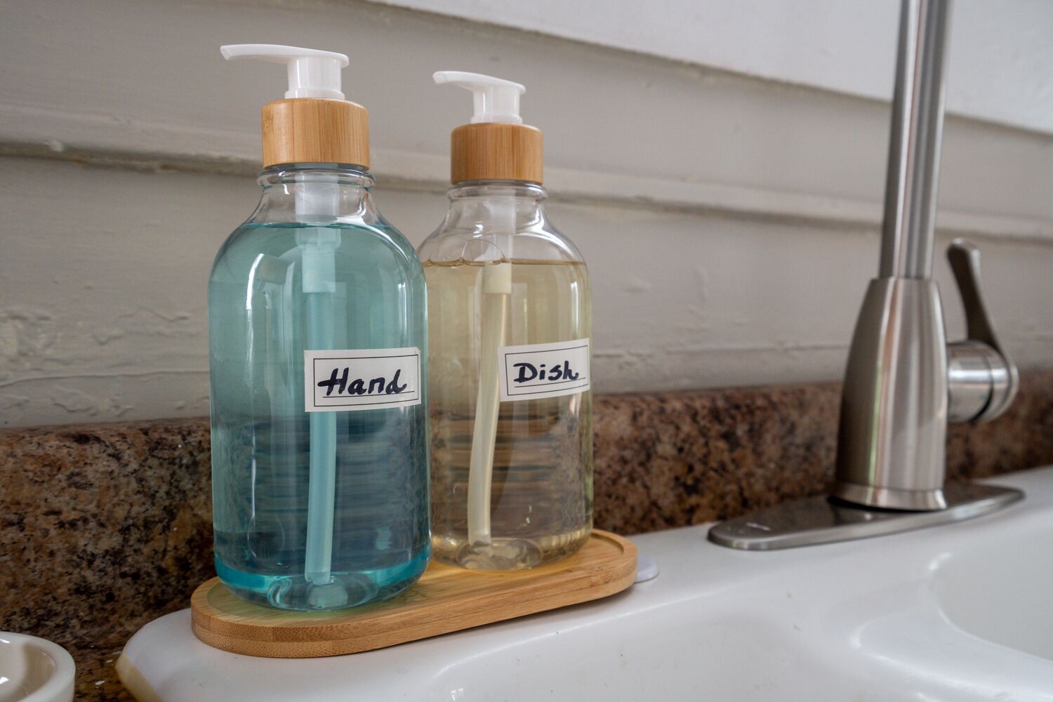 Zwei Glasflaschen mit Pumpspender auf einem Holzuntersetzer, beschriftet mit „Hand“ und „Geschirr“, auf einem Badezimmerwaschbecken.