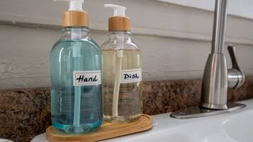 Zwei Glasflaschen mit Pumpspender auf einem Holzuntersetzer, beschriftet mit „Hand“ und „Geschirr“, auf einem Badezimmerwaschbecken.