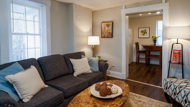 A living room with a gray sofa, a round coffee table with decorative items, a dining area with a wooden table and chairs, and a kitchen visible in the background. - *5 Min to WVU*Smart TV*2 Full Kitchen*W/D (Morgantown)