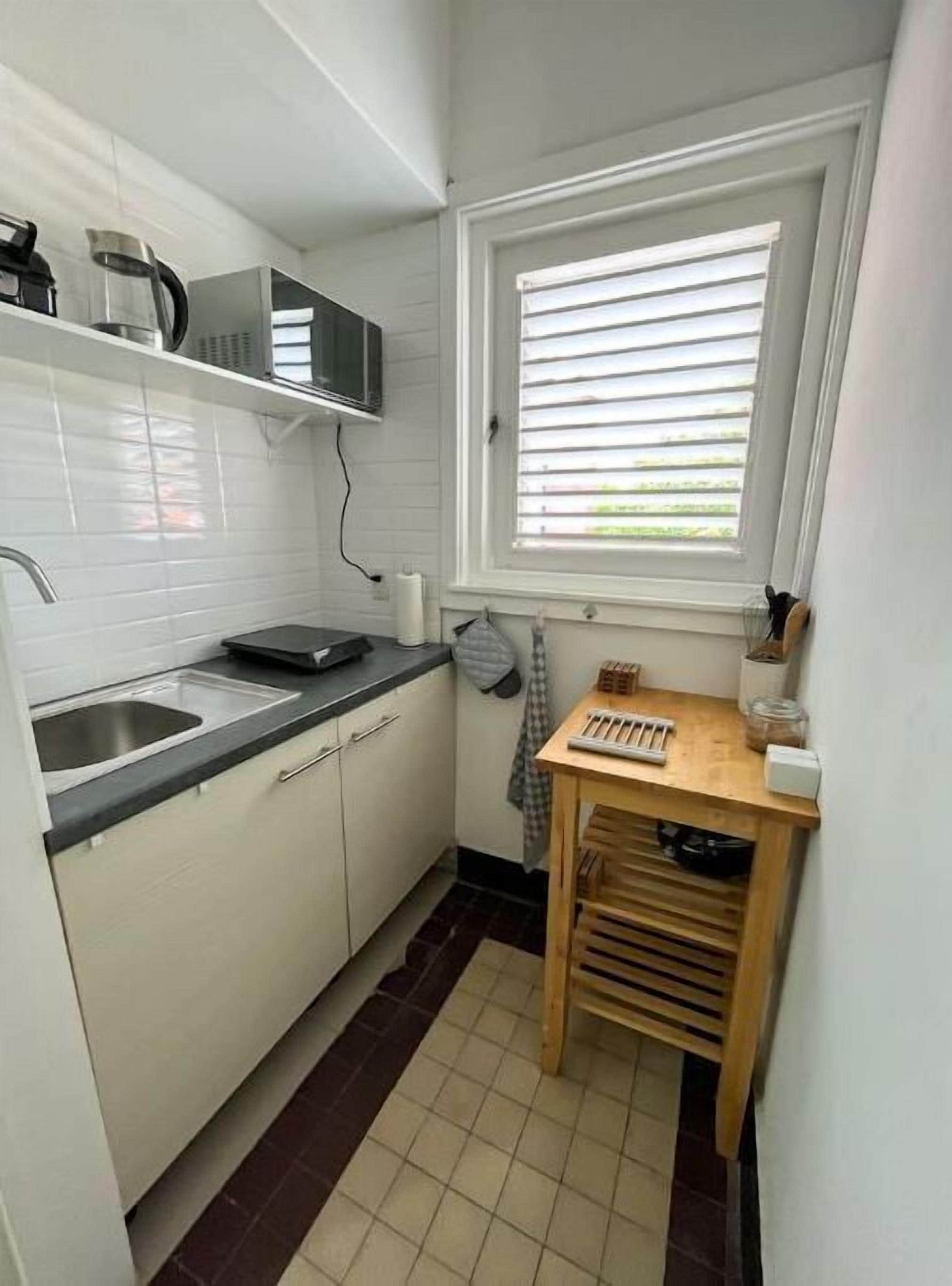 Eine kleine Küche mit weißen Schränken, einer Edelstahlspüle, einer Mikrowelle und einem Fenster mit Jalousien.