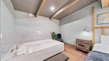 Ein modernes Badezimmer mit einer großen Badewanne, einer Holzbank, einem kleinen Nachttisch und einem Bett.