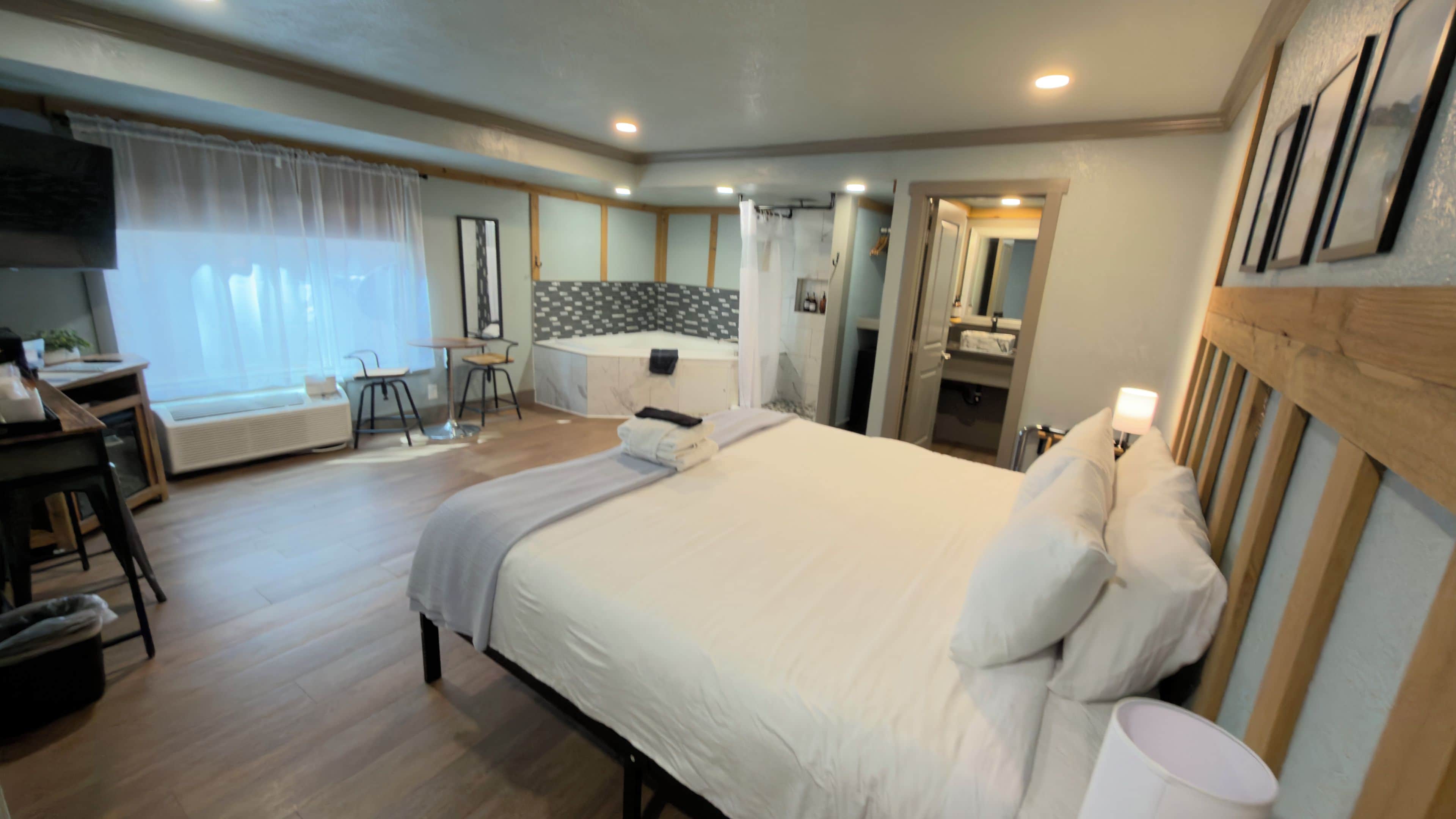 A hotel room with a bed, a desk, a chair, a small table, a mirror, and a window with curtains.