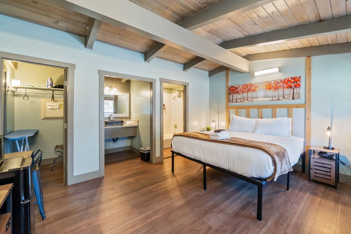 A modern bedroom with a wooden floor, a bed with white linens, a nightstand, a bedside table, a potted plant, and a painting on the wall.