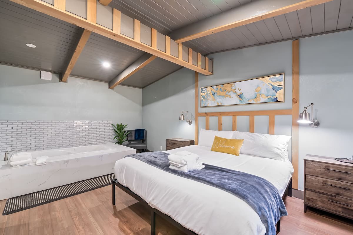 A modern bedroom with a large bed, a built-in bathtub, wooden beams, a map artwork, bedside lamps, and a bedside phone.