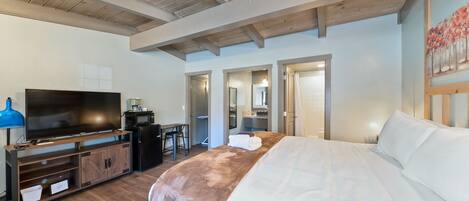 A modern hotel room with a large bed, a flat-screen TV, a kitchenette, and a bathroom visible through an open door.