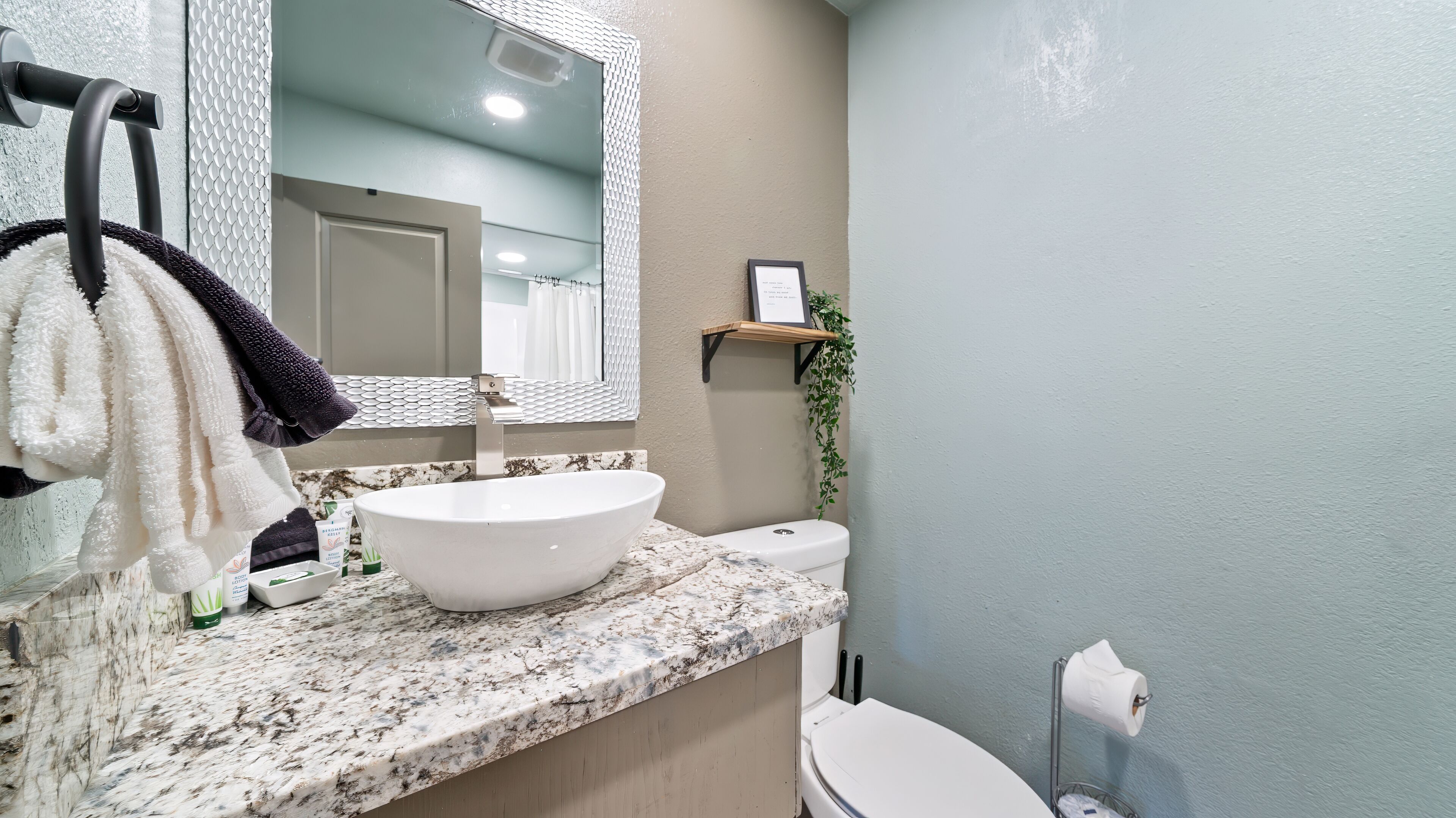 Une salle de bain avec un lavabo blanc, un comptoir en marbre, un porte-serviettes avec des serviettes, un certificat encadré posé sur une étagère et des toilettes.