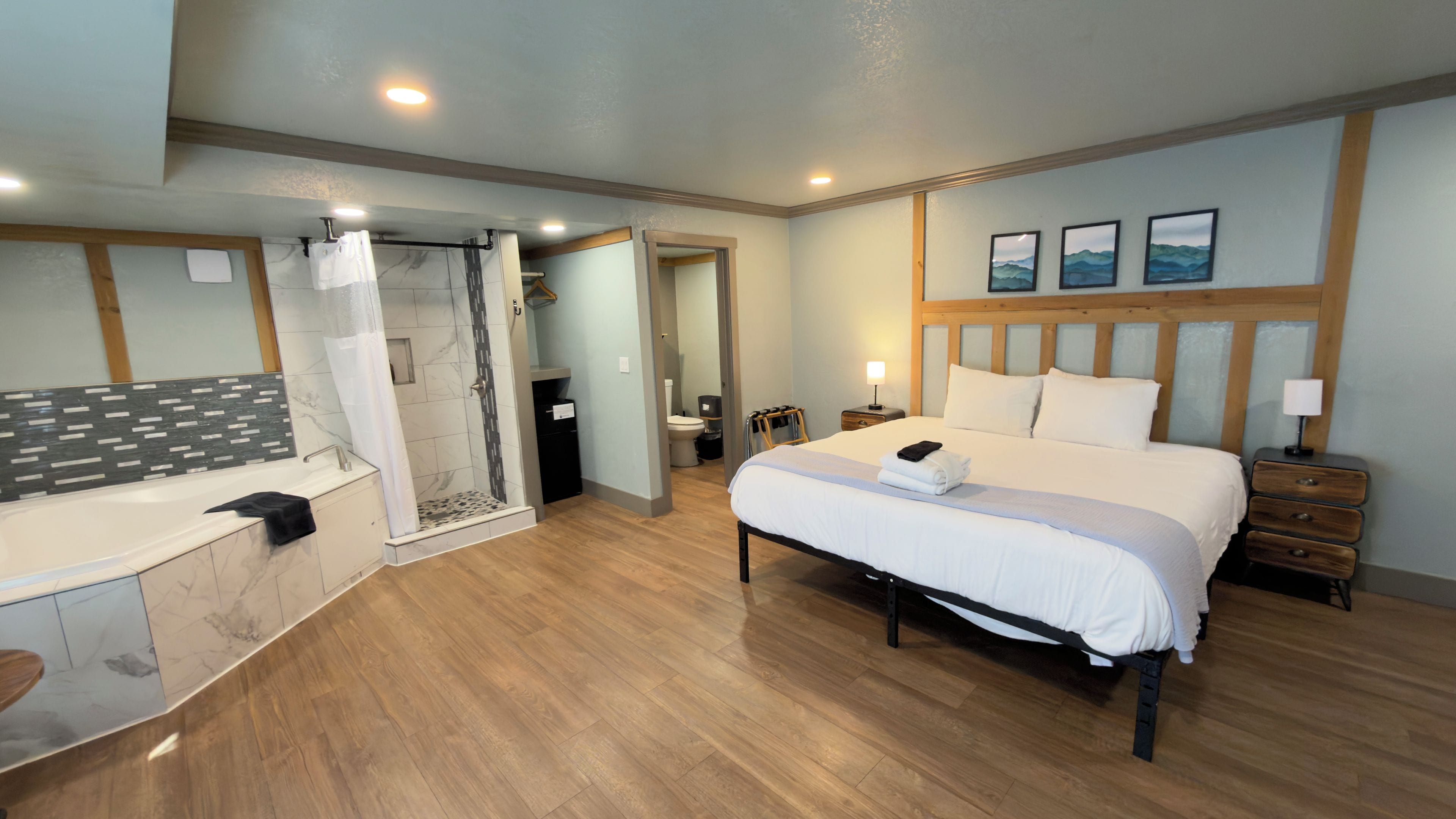 A hotel room with a large bed, a shower area, a vanity with a mirror, and a wooden floor.