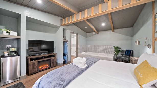 A modern bedroom with a large bed, a television, a fireplace, and a bathroom with a white brick wall. - Blue Jay Lodge (South Lake Tahoe)