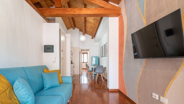 A modern living room with a blue sofa, wooden ceiling beams, a flat-screen TV mounted on the wall, and a dining area in the background. - La Terrazza della Marina (Cagliari)