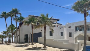 Un edificio blanco con techo plano y puerta de garaje, rodeado de palmeras y arena.