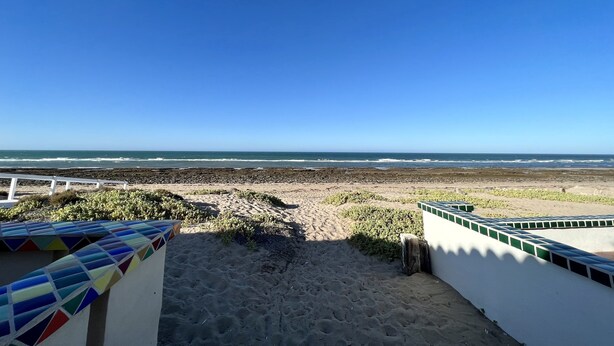 Vista de la playa con un sendero arenoso que conduce al océano, una barandilla blanca y una estructura de azulejos coloridos.