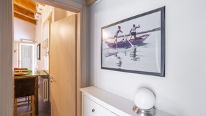 A room with a wooden ceiling, a white wall, a framed photograph of two people rowing a boat, a white cabinet with a spherical object on top, and a door leading to another room.