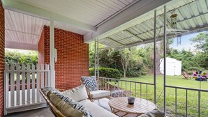Un porche techado con muebles de mimbre, una mesita y vistas a un jardín y un cobertizo.
