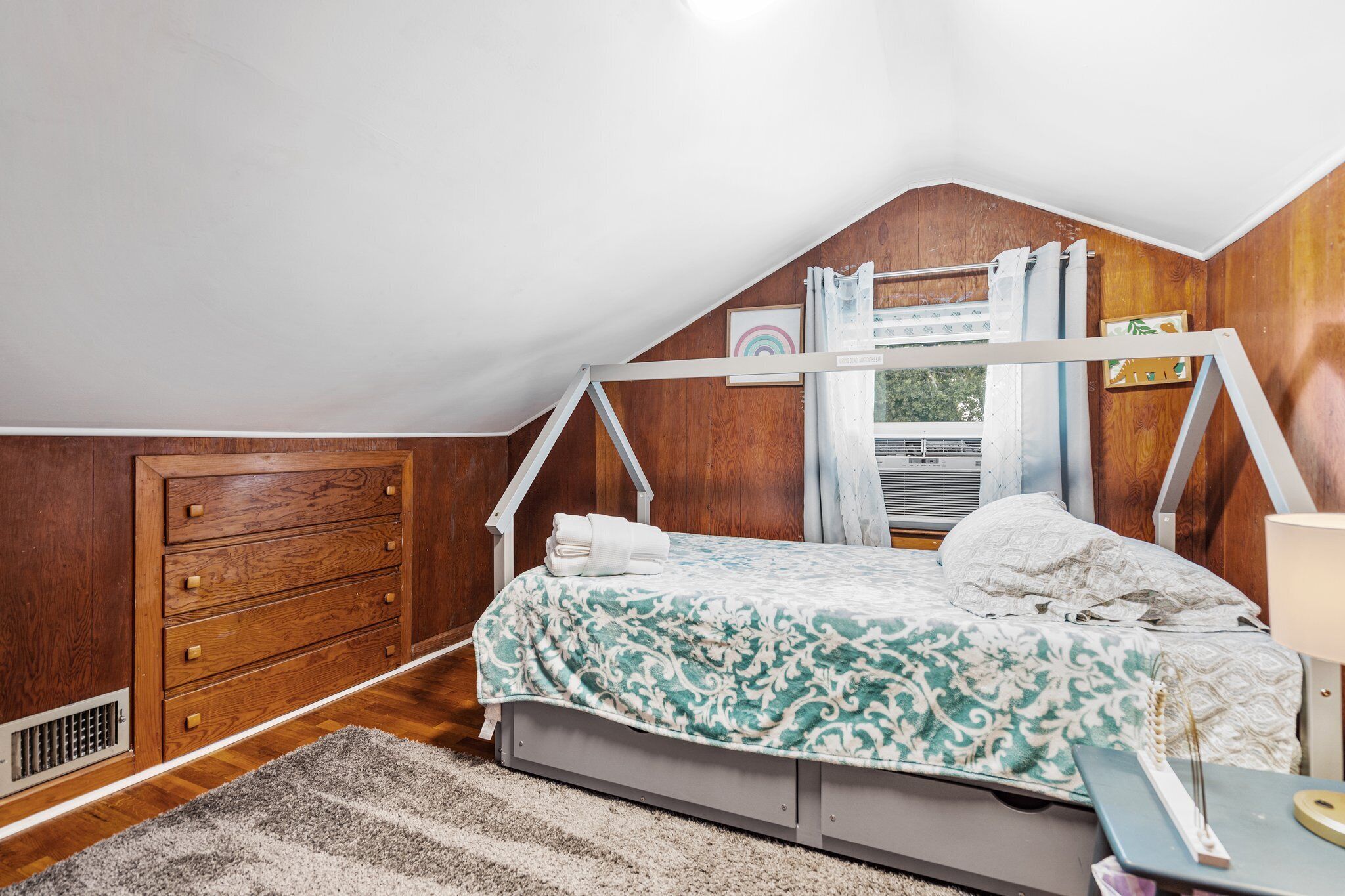 A bedroom with a sloped ceiling, wooden walls, a bed with a patterned bedspread, a bedside table, and a window with curtains.