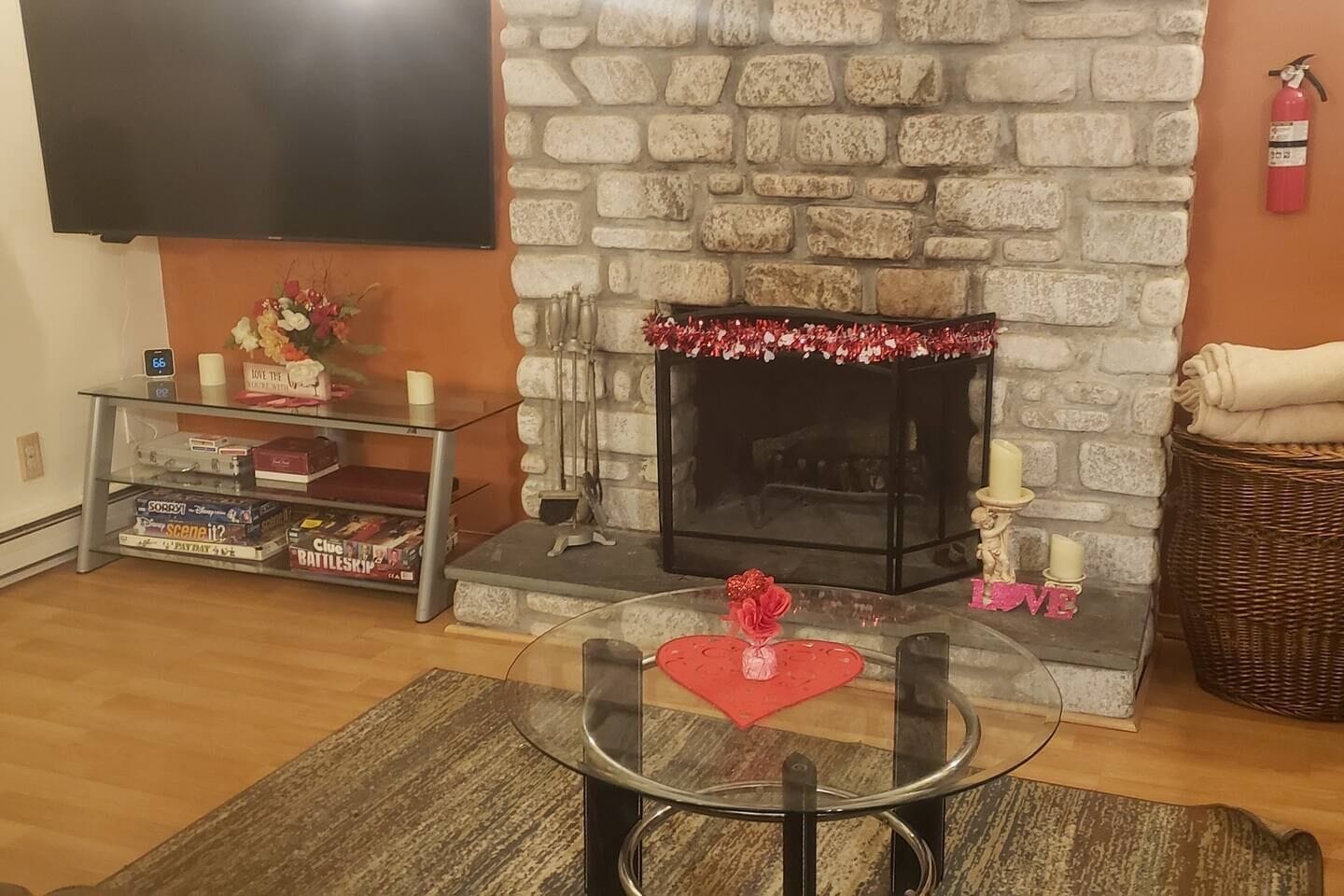 A living room with a stone fireplace, a flat-screen TV mounted on the wall, a glass coffee table with a red heart decoration, and a wicker basket.